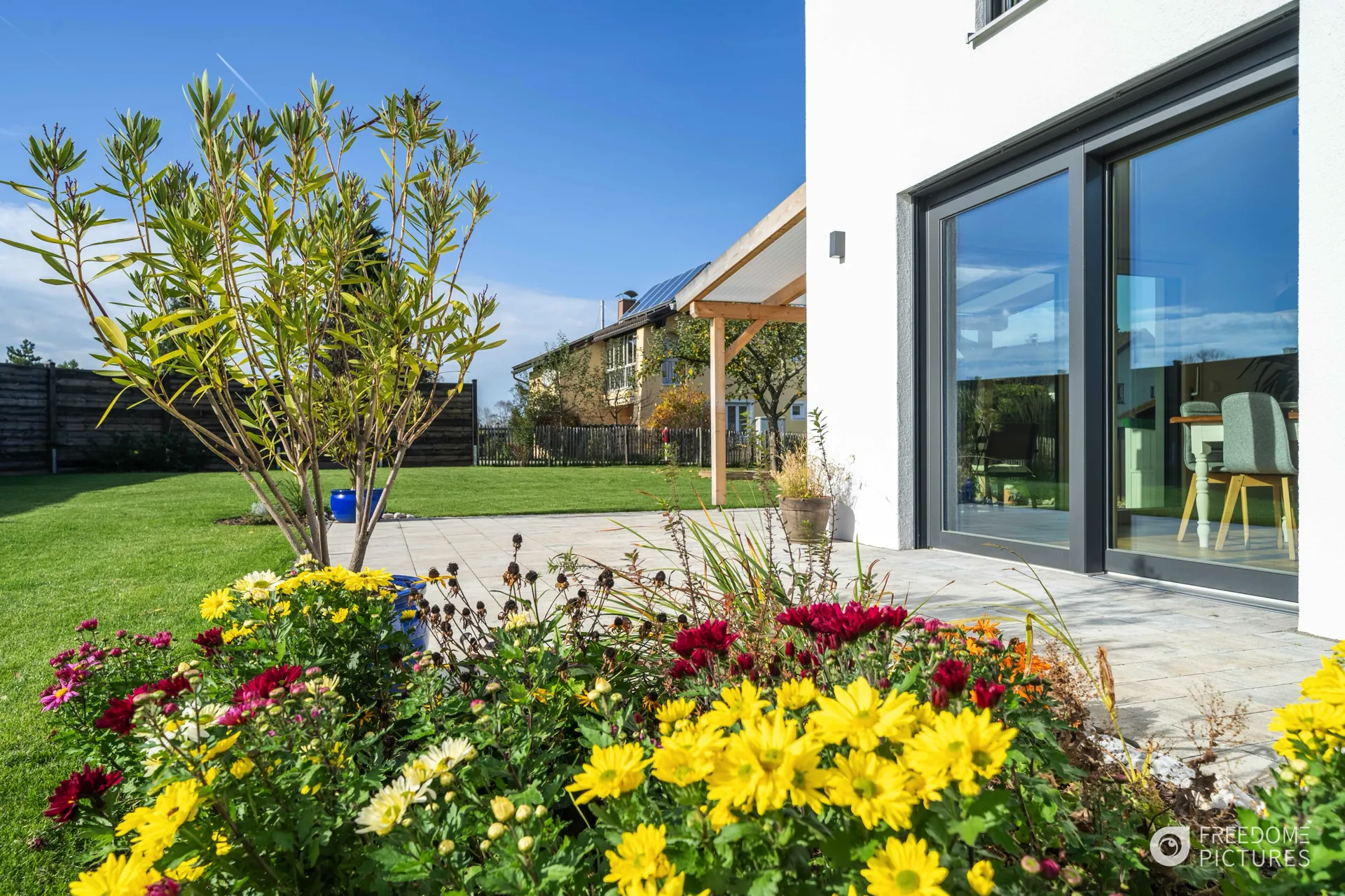 Sonniger Blick über bunte Blumen im Vordergrund auf eine Haus-Terrasse und frisch gemähte Wiese im Garten