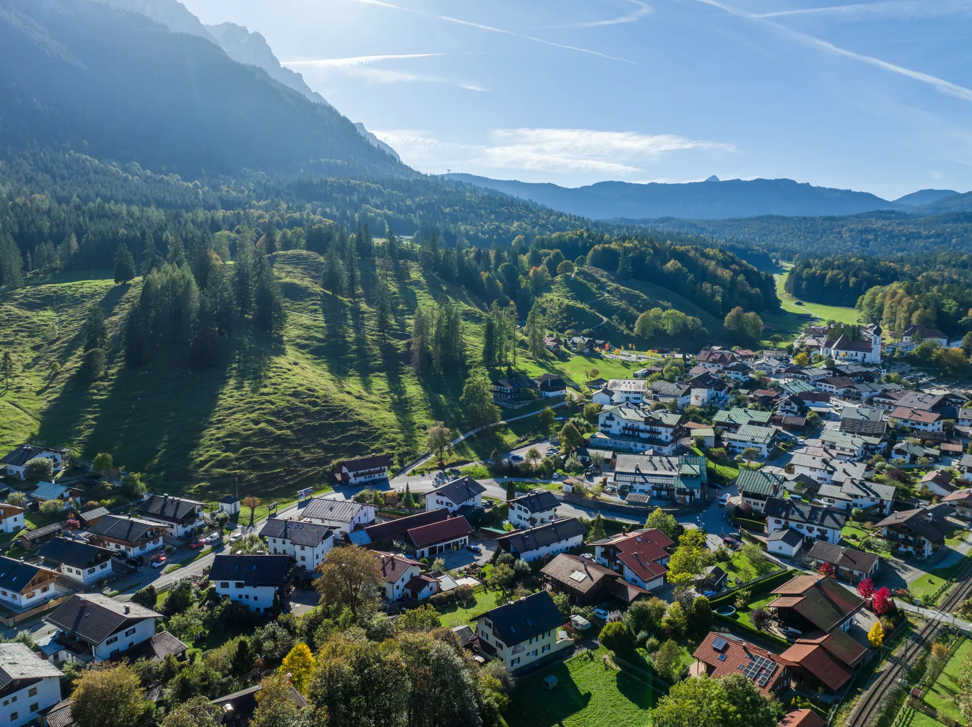 Atmosphärische Drohnenaufnahme eines Wohngebiets in Garmisch-Partenkirchen mit Alpenpanorama im Hintergrund und sonnigem Gegenlicht